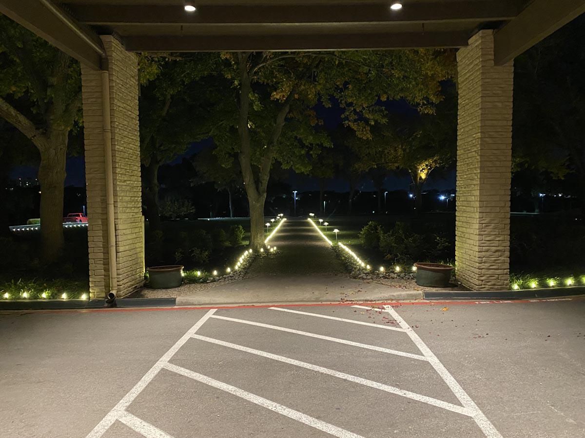 Entrance with walkway, flanked by columns, lined with lights, and leading to a tree-lined area at night.