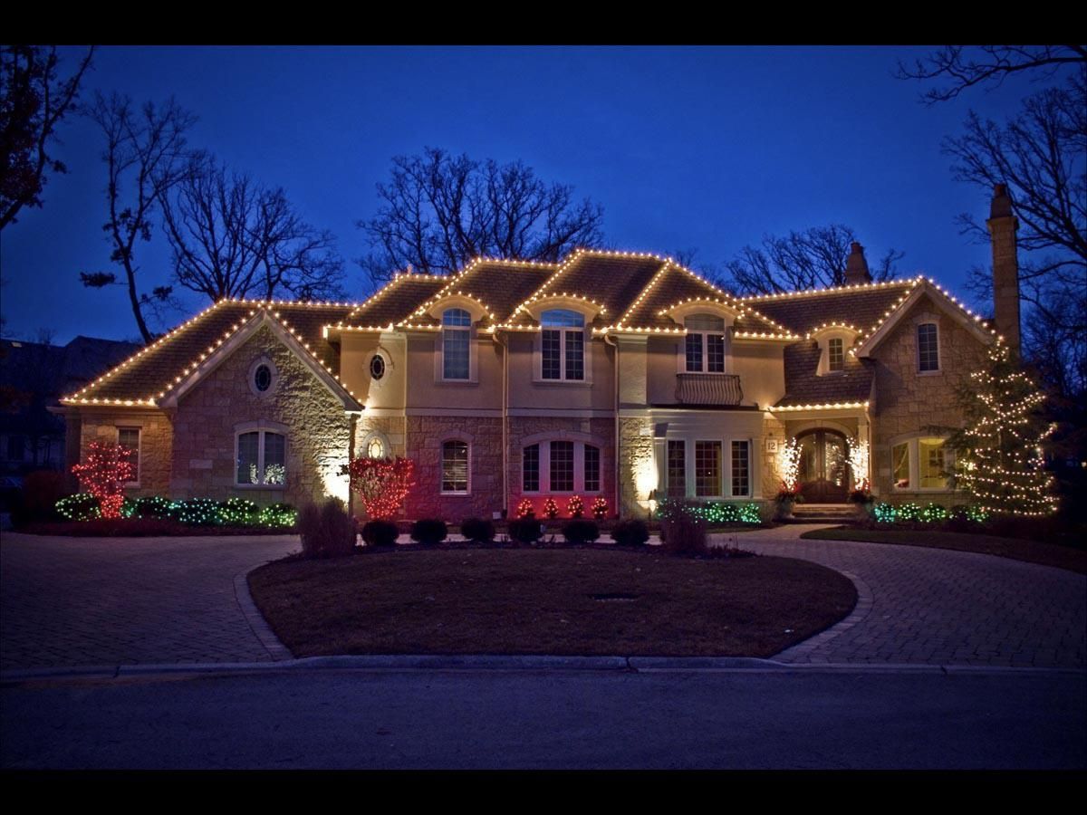 House decorated with white Christmas lights on the roof and colored lights on the ground, at dusk.