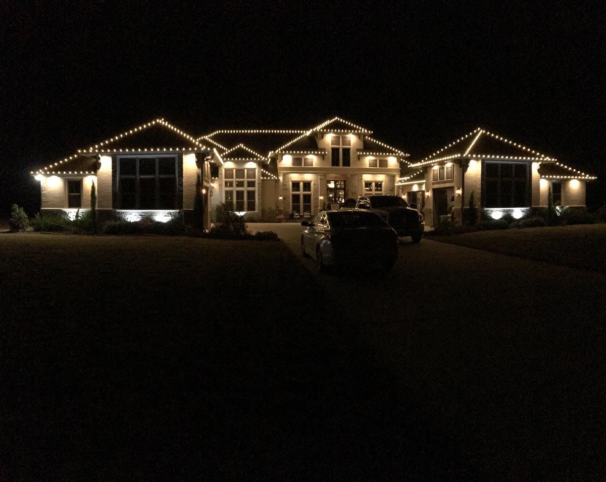 House at night, illuminated with white lights along rooflines and spotlights; cars in driveway.
