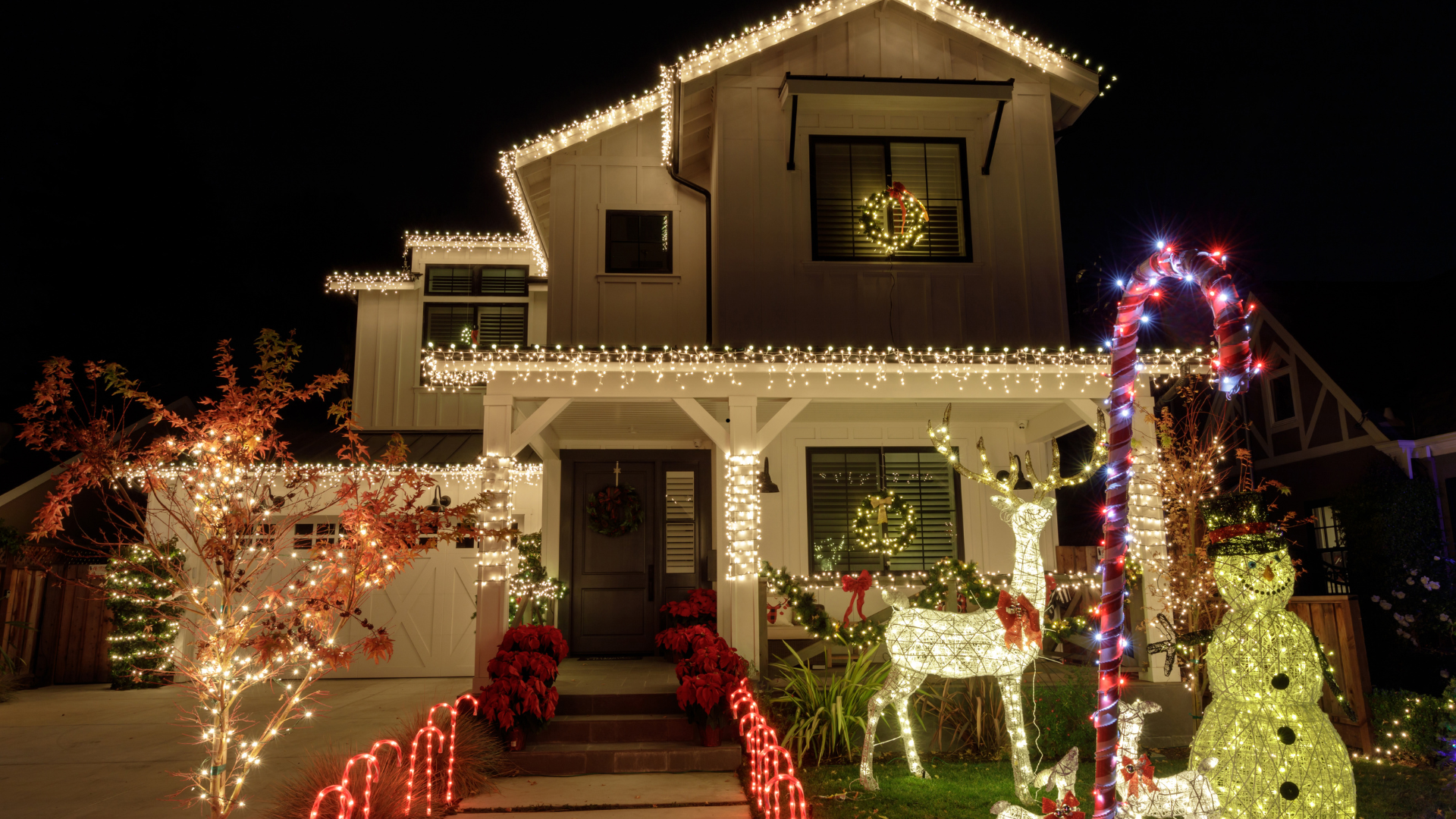 Festive house with white siding and Christmas lights, featuring reindeer and snowman lawn decorations.