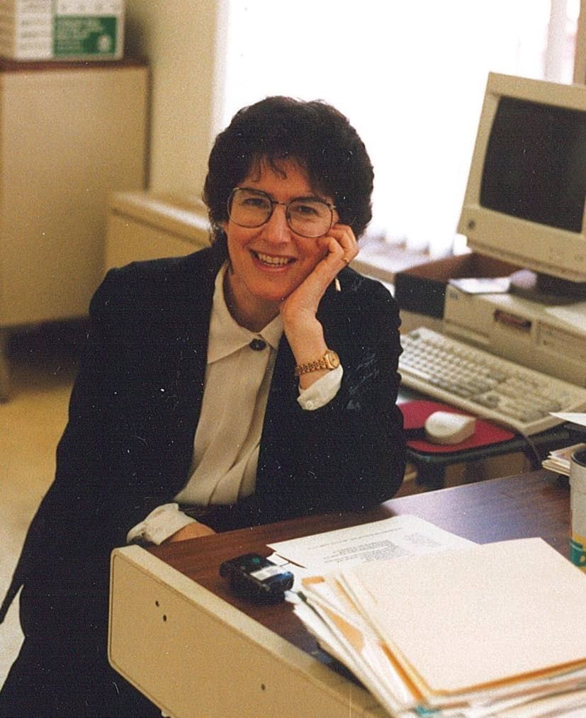 Sally Borden sits in the KidSafe office in a black blazer and white shirt, it is 1998 and the computer next to her reflects this 