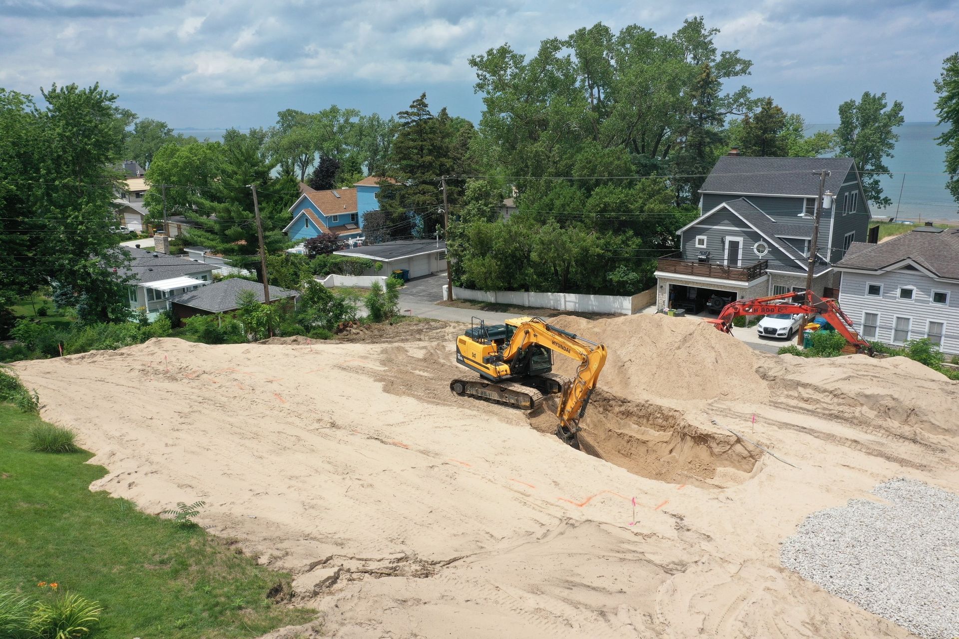 Excavator digging on a sandy lot near houses and a lake on a sunny day.