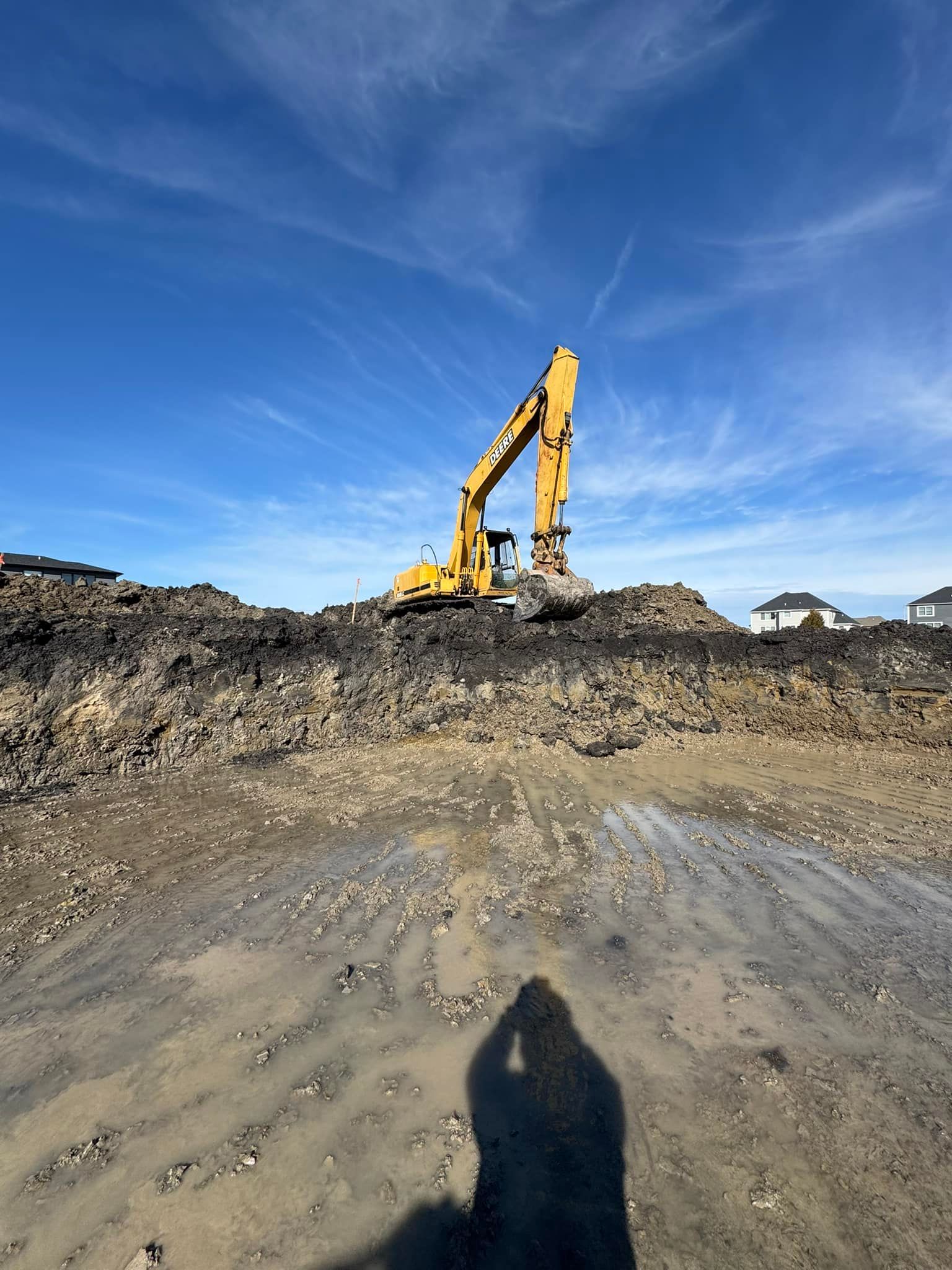 Yellow excavator on dirt mound under a blue sky, shadow of person in foreground.