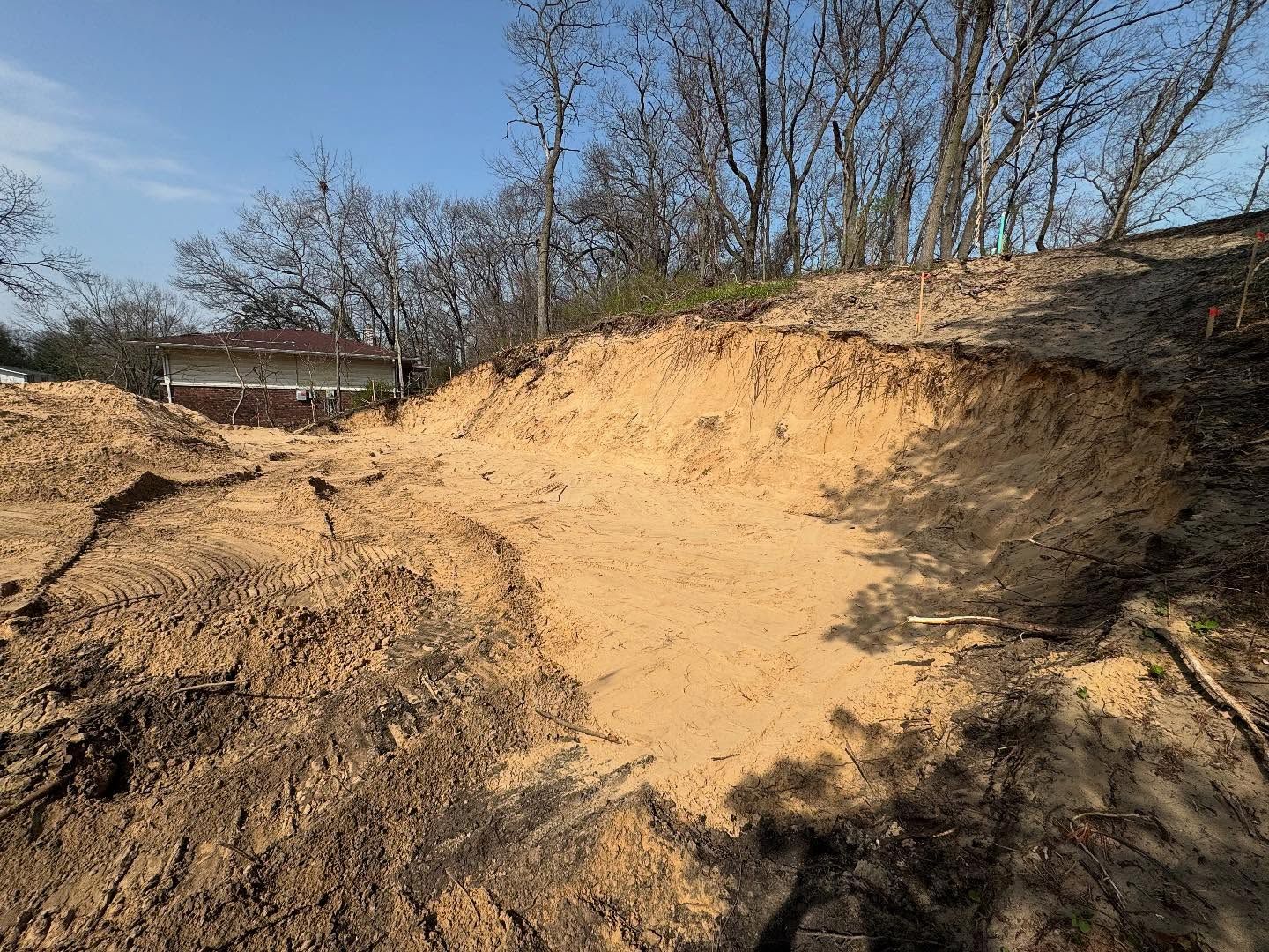 Erosion exposing sandy soil in a clearing with trees and a distant house under a blue sky.