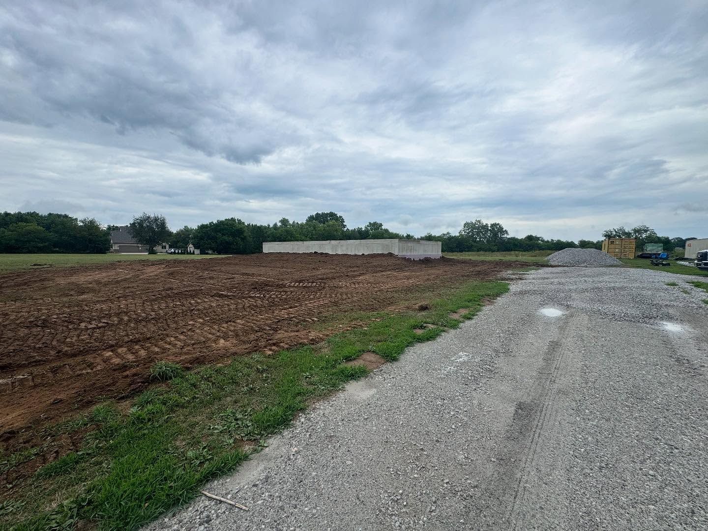 A construction site under a cloudy sky. Brown, plowed earth, a gravel road, and a concrete foundation are visible.