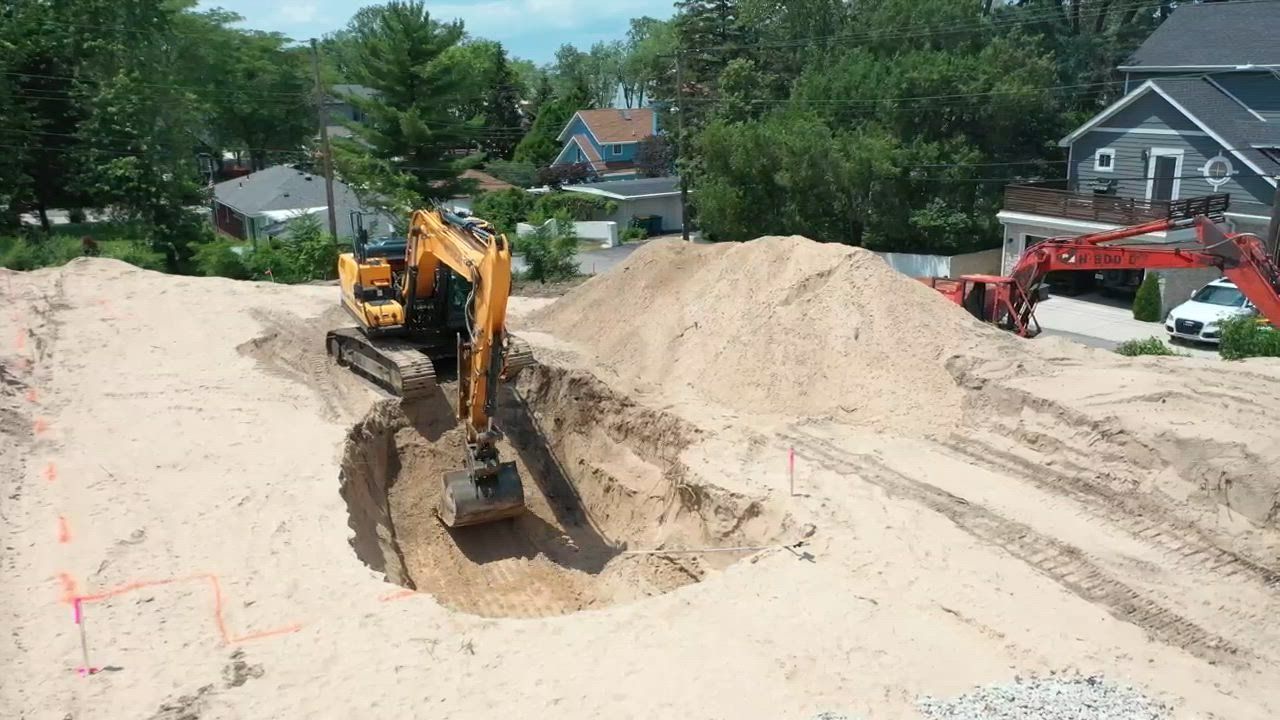 Yellow excavator digging a foundation pit on a construction site. Dirt piles and houses in background.