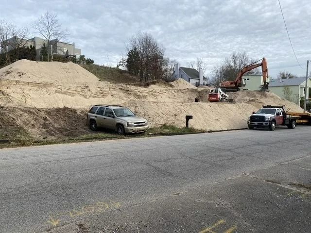 Construction site with large piles of wood chips. An SUV and truck are parked on a street. An excavator and skid steer are working.