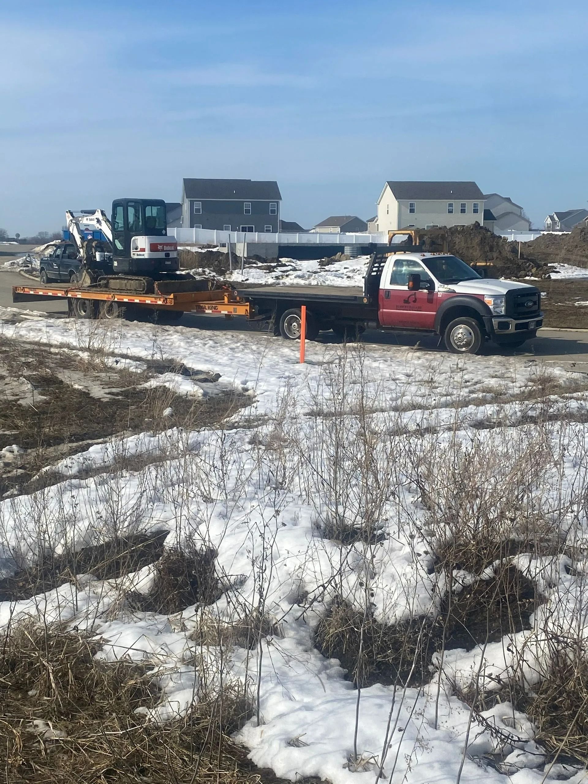 Construction site with a truck, trailer, and excavator on snowy ground with houses in the background.