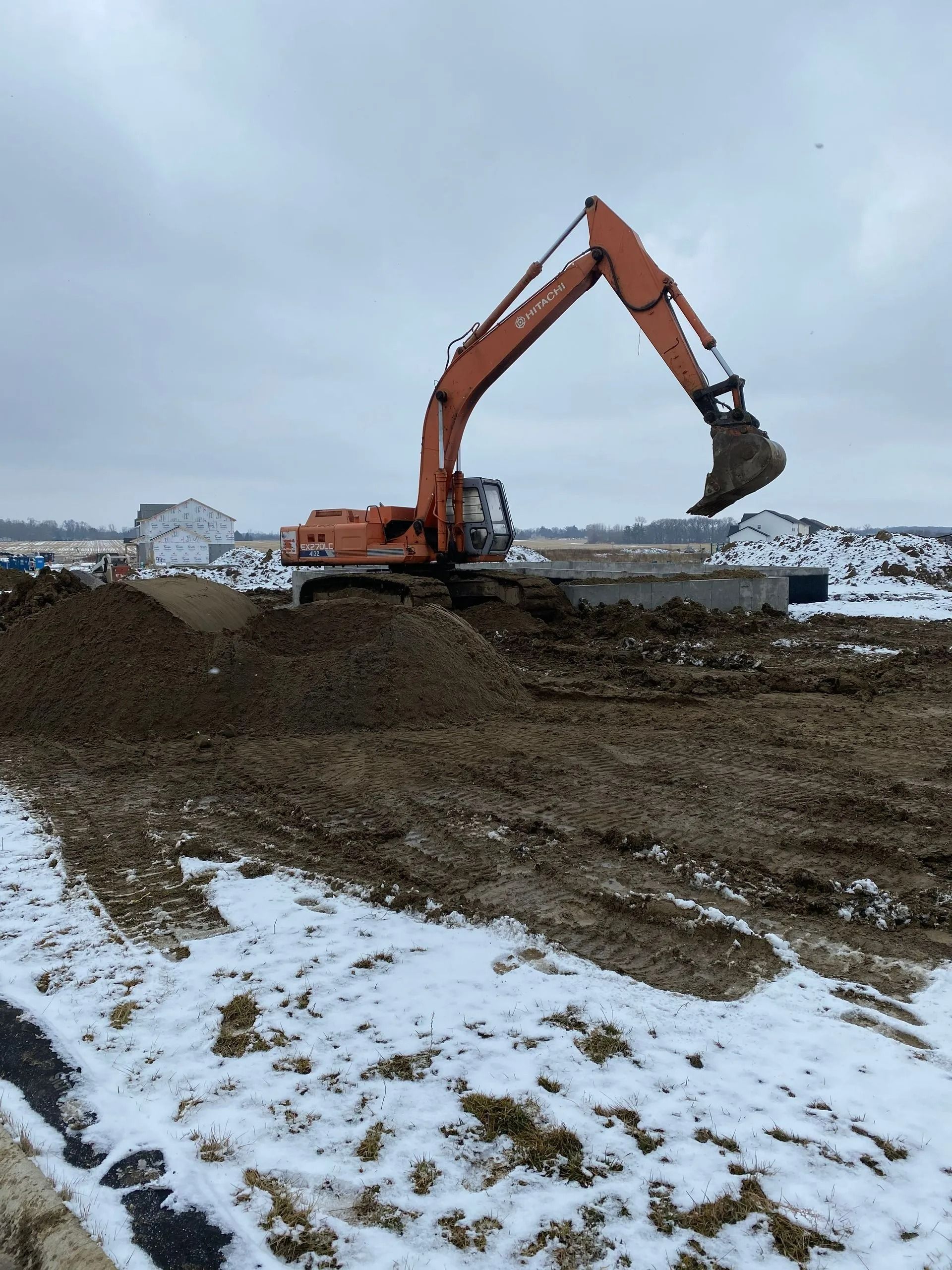 Orange excavator moving dirt on a construction site, with snow and cloudy sky in the background.