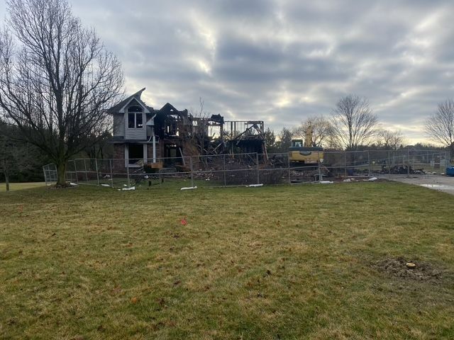 House destroyed by fire, surrounded by fencing. Burned structure against cloudy sky. Yellow construction vehicle visible.