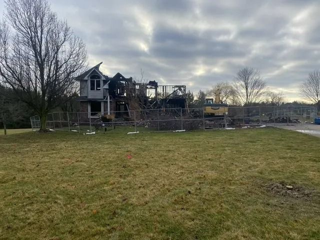 House destroyed by fire, surrounded by fencing, construction equipment visible. Cloudy sky.
