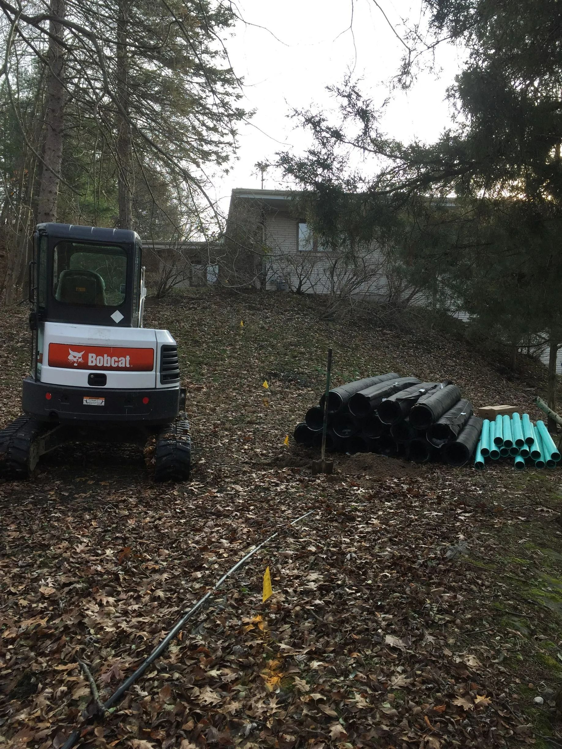 Bobcat excavator on a leaf-covered hillside next to a pile of black and green pipes, with a house in the background.