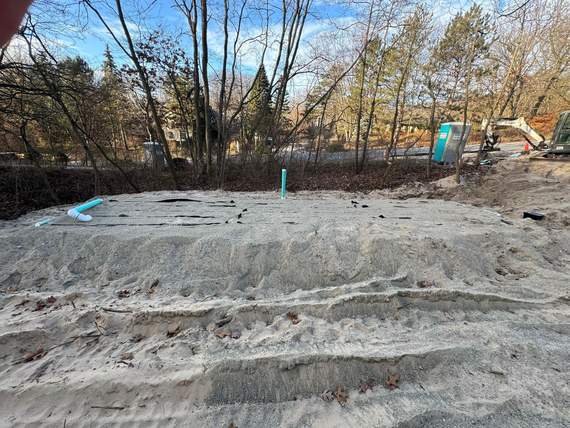 A sand-covered construction site with exposed pipes. Trees and equipment are visible in the background.
