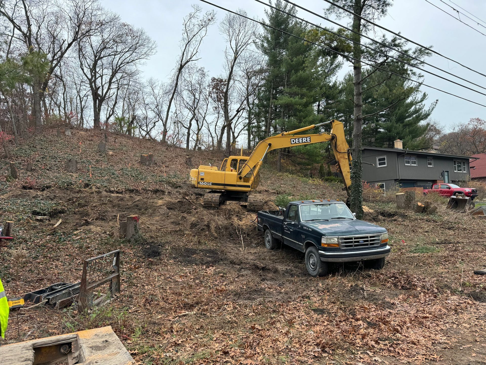 Yellow excavator clearing brush from a hillside, with a blue pickup truck in front of it.