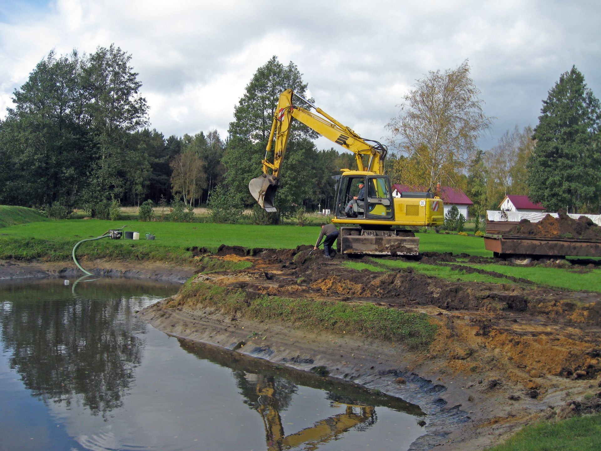 Yellow excavator dredging a pond's edge, grassy bank, trees, and sky reflecting in the water.