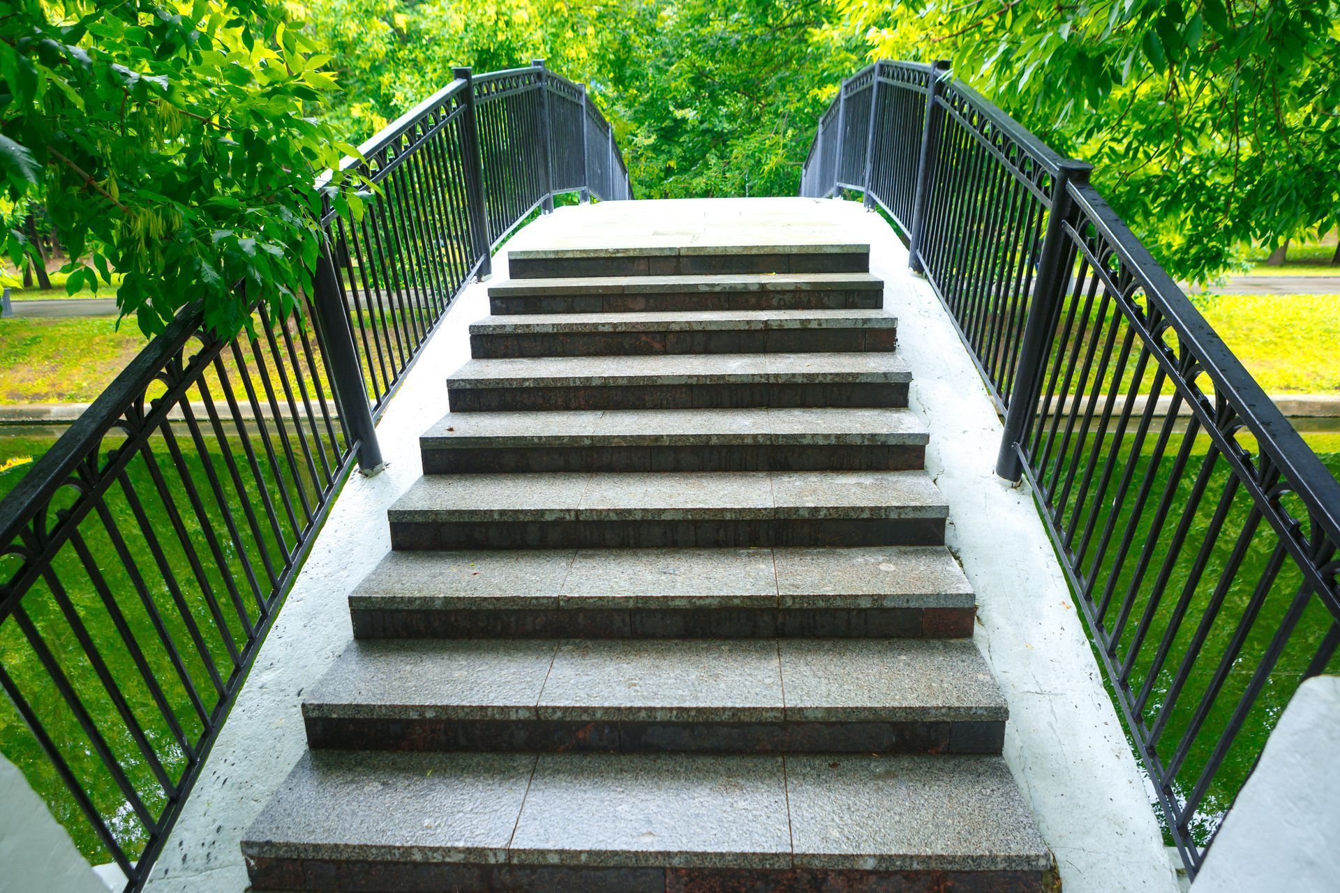 Stone steps leading up a pedestrian bridge with black railings are surrounded by green trees Stone steps leading up a pedestrian bridge with black railings are surrounded by green trees