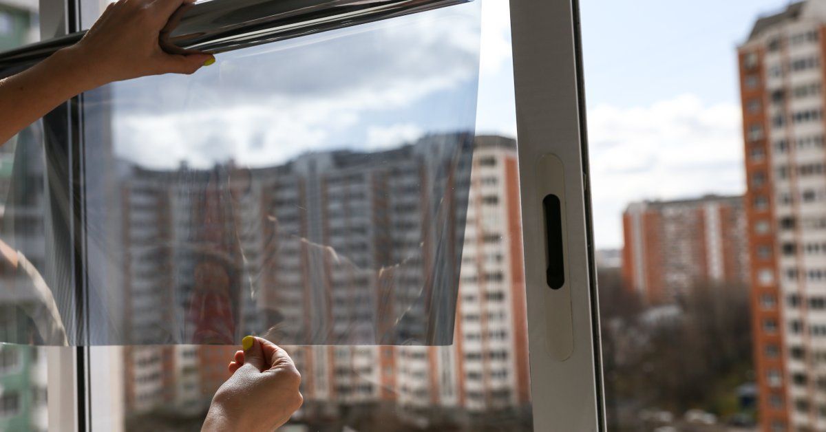 A pair of hands with yellow painted nails unrolling a film of window tint. Outside the window are apartment buildings.