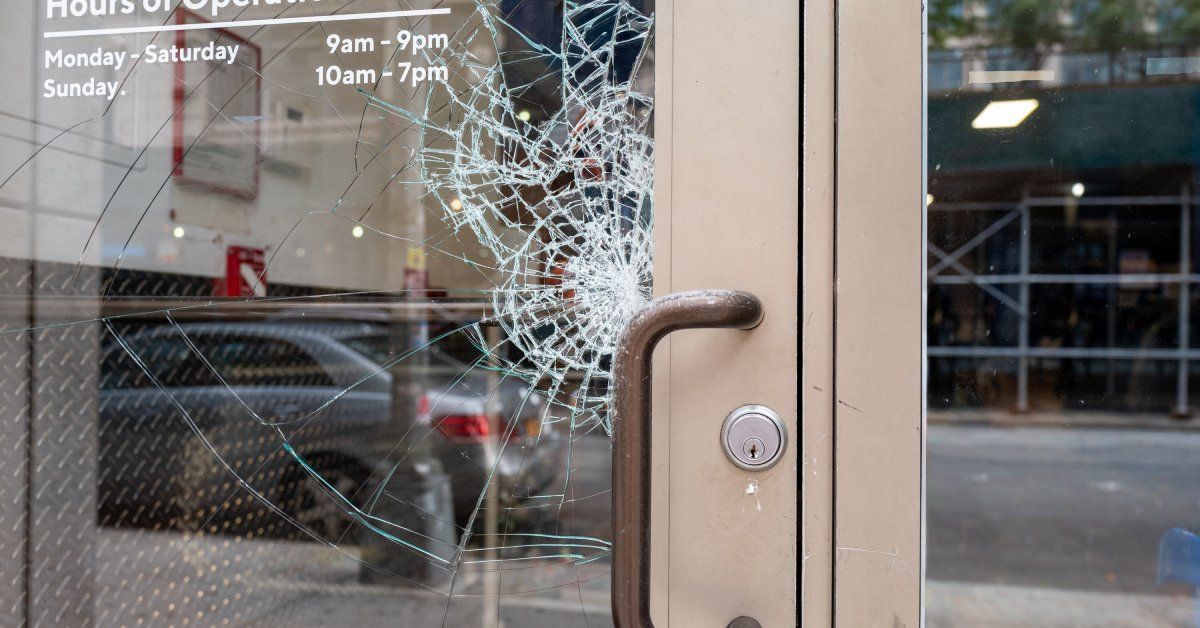 The front door of a business with the glass broken by the handle. The hours of operation are posted on the glass.