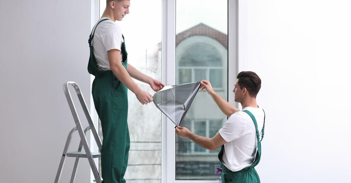 Two workers in white T-shirts and green overalls are rolling out a sheet of window film. One is on a ladder.