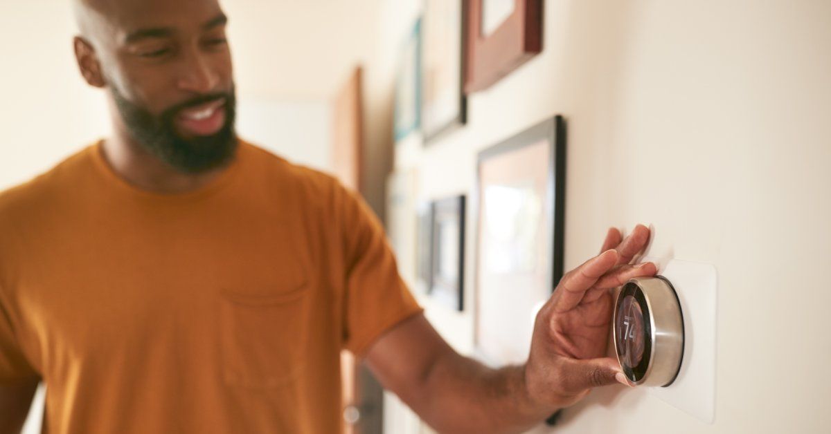 A man with a beard, wearing a yellow shirt, reaching out to touch the circular, wall-mounted thermostat.