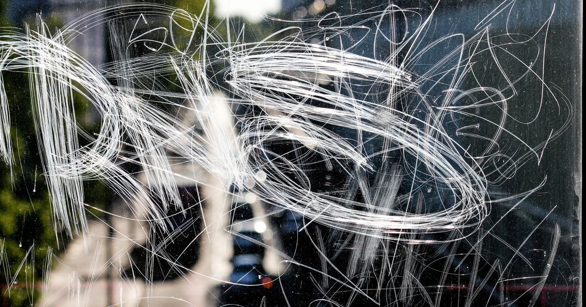 A close-up shot of a window pane covered in scratches of graffiti in different letters. The window reflects a busy road.