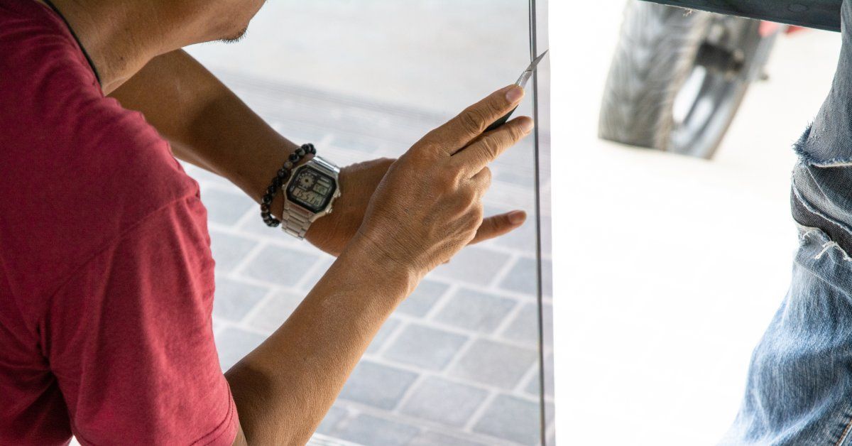 A focus shot of a man in a red t-shirt holding a sharp blade to trim the edges of a window tint.