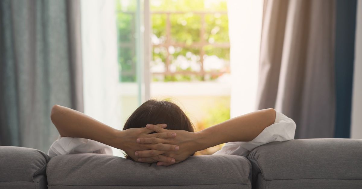 The back view of a grey couch and a woman relaxing on it with her hands behind her head. The couch is in front of a window.