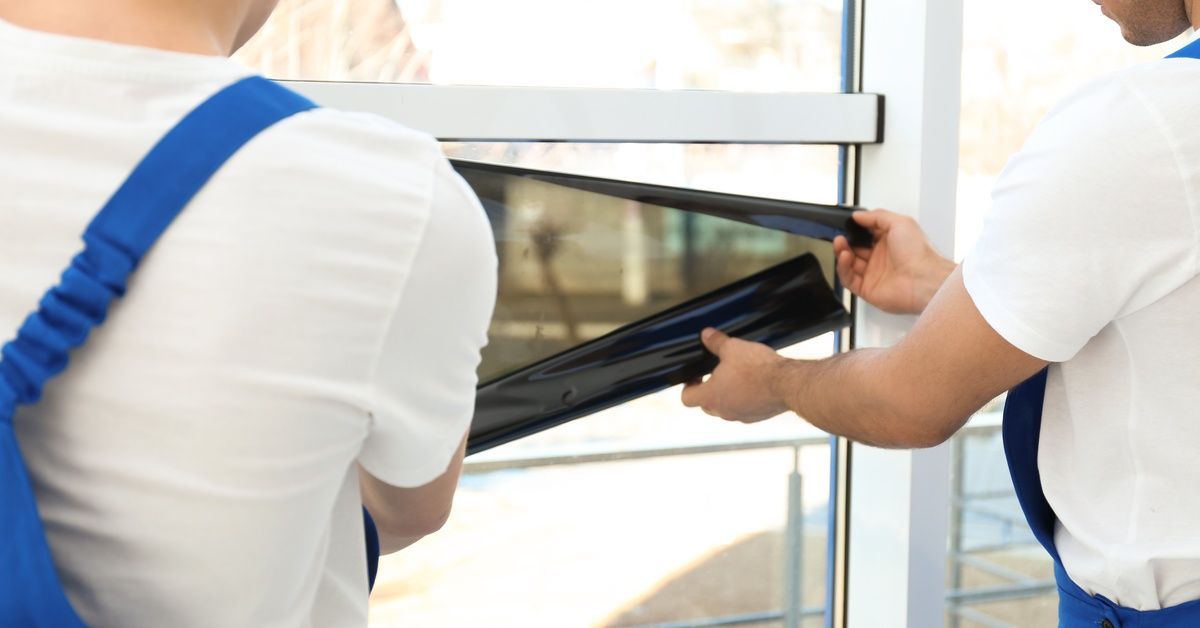 Two professional workers in white t-shirts and blue overalls unrolling a dark sheet of window film.