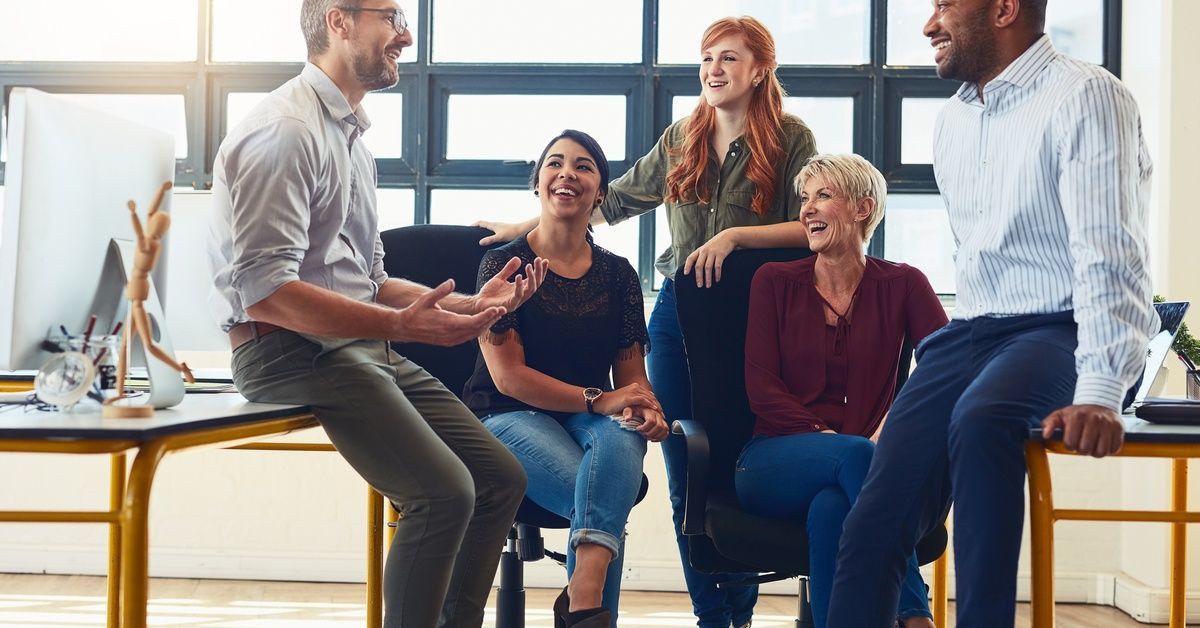 A group of five workers in casual work attire, sitting in a group, gesturing and smiling as they talk.