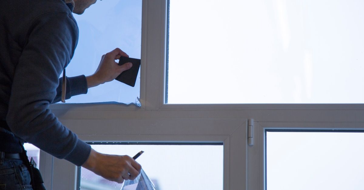 A man in a hoodie using a card to press down dark window film on a set of windows to remove any bubbles.