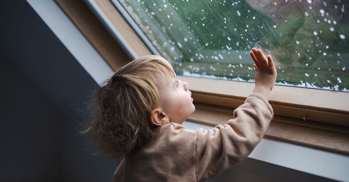 The toddler is inside the home with the raindrops on the outside of the window.