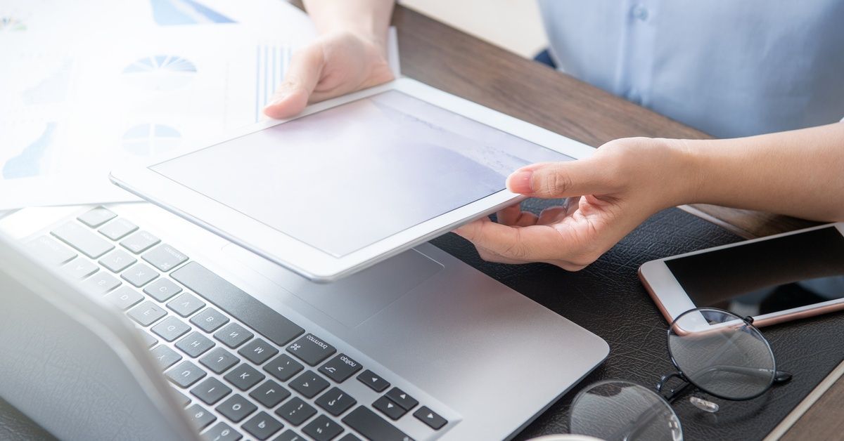 Bright window glare washes over a white tablet, a silver laptop, and a small stack of papers on a modern office desk.