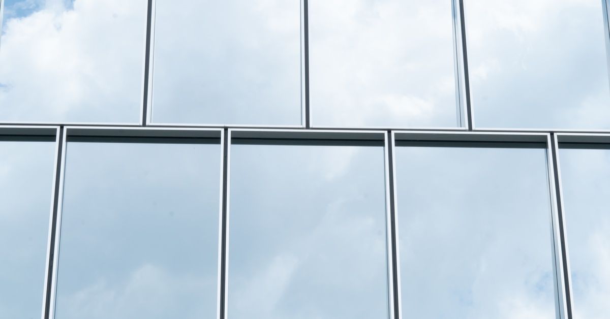 Two rows of reflective glass windowpanes on an office building mirror a blue sky and white clouds.