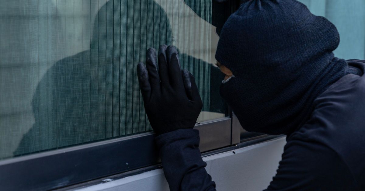A burglar stands near a window at night, trying to look in, but the blinds are closed.