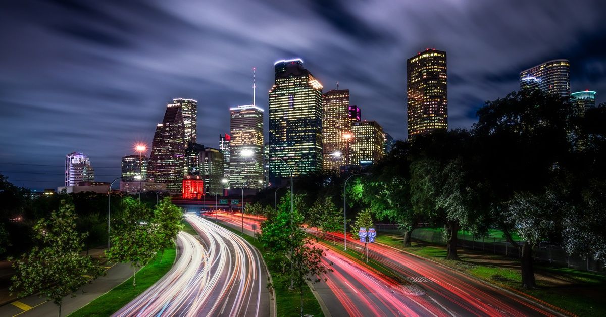 A nighttime skyline view of Houston with highways below, where red and white car lights blur together in a time-lapse effect.