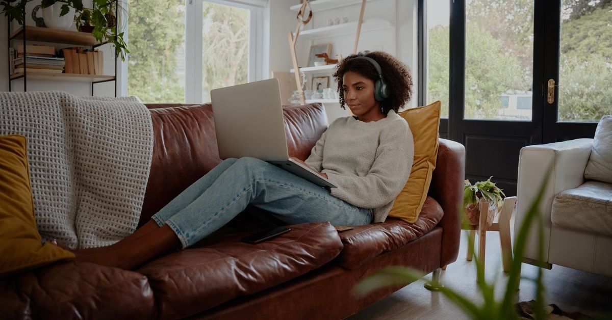 A young woman wearing headphones sits on her couch using a laptop, with large windows behind her in the living room.