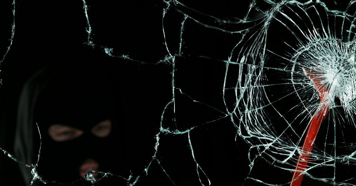 A masked burglar strikes a window with a red crowbar, but the glass remains intact during the attemp