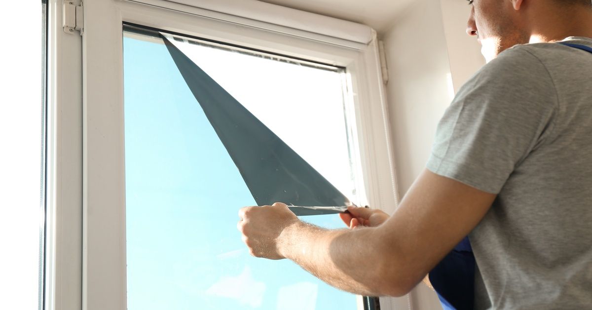 A man in a white T-shirt and blue coveralls peels back a corner of the window tint film to inspect and repair it.