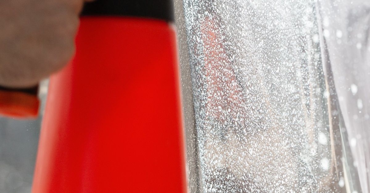 A person sprays window tint film with a red water mister while making repairs on a window in bright daylight.