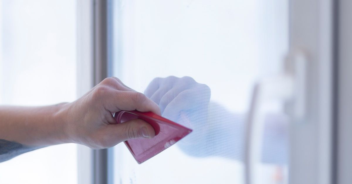 A person uses a red plastic scraper to remove bubbles from under the window tint film on a window.