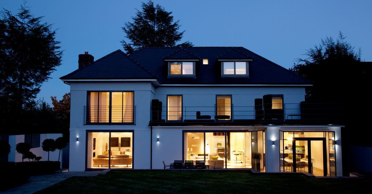 A two-story white modern home at night shows brightly lit interiors clearly visible through expansive windows.