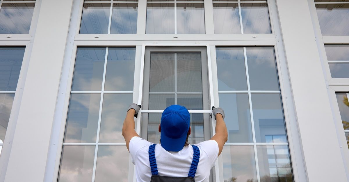 A man installs mesh solar screens on tall exterior windows of a large white-framed home while wearing gloves and a hat.