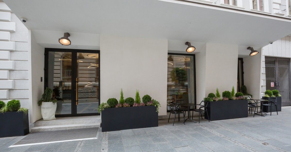 A modern white building has a glass-front entrance and windows overlooking patio tables and chairs o