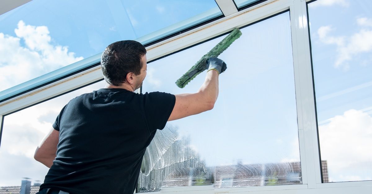 Man cleaning large glass windows with a long squeegee, removing soap and water under a bright blue sky.