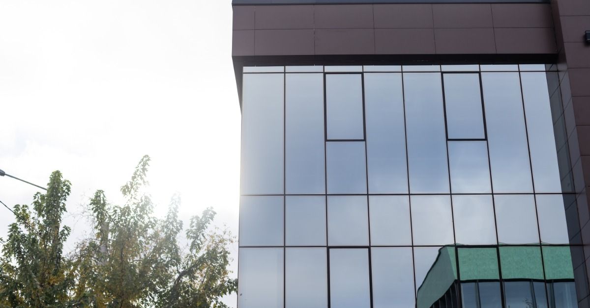 A modern building with large reflective glass panels stands beside leafy trees under a clear sky.