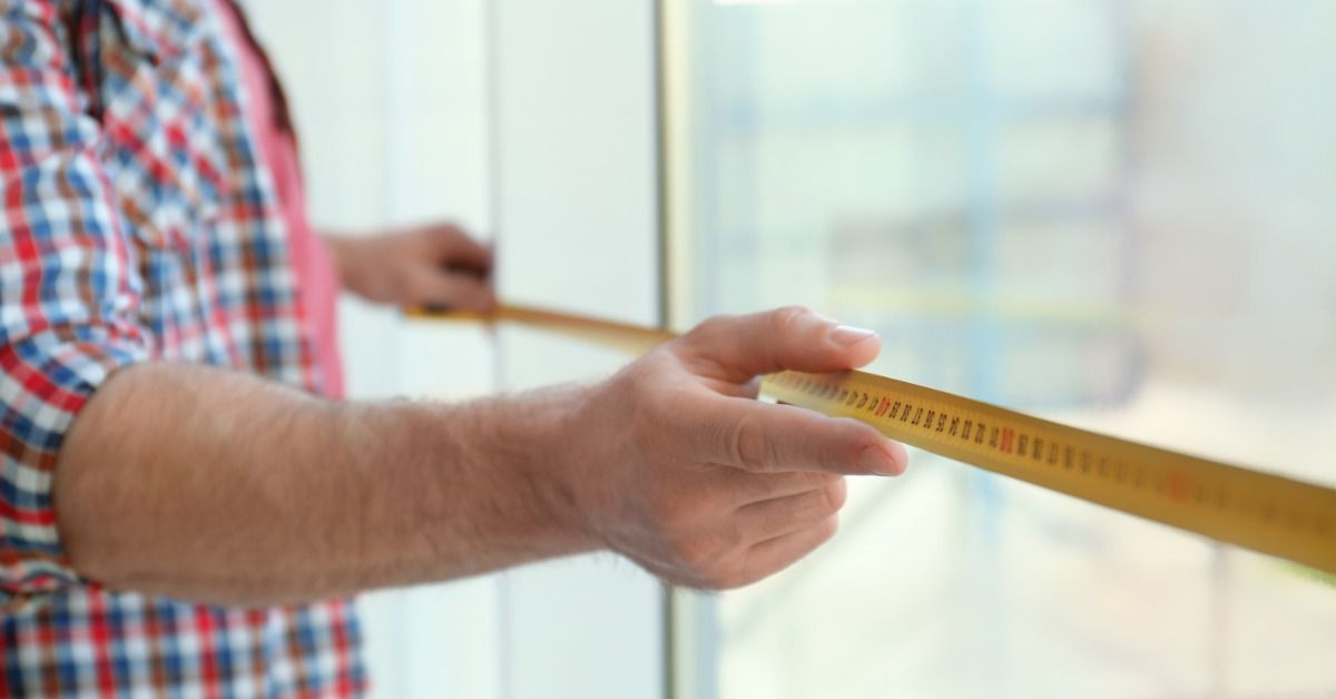A person measures a window frame with a tape measure, holding it steady while checking dimensions indoors.