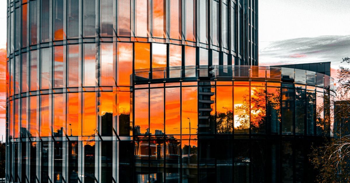 A curved glass office building is reflecting a vivid orange sunset across its modern facade and skyline.