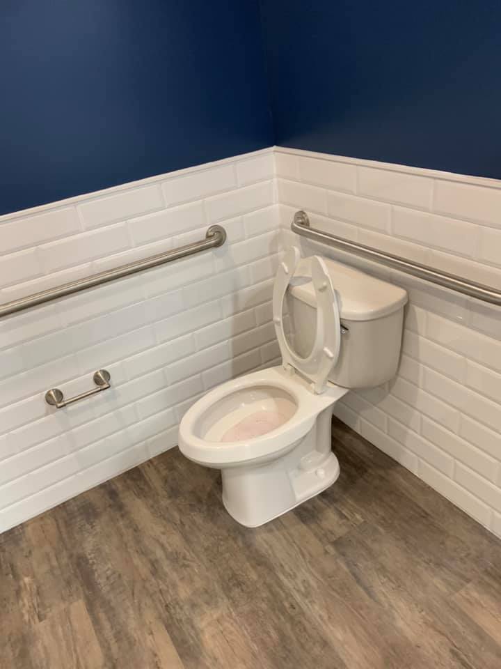 Toilet in a corner of a bathroom with grab bars on white tiled walls and wood-look flooring.