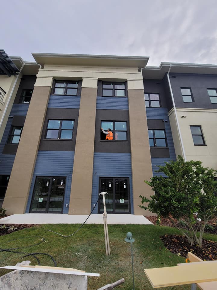 A person in an orange vest works on a window of a multi-story apartment building under a cloudy sky.