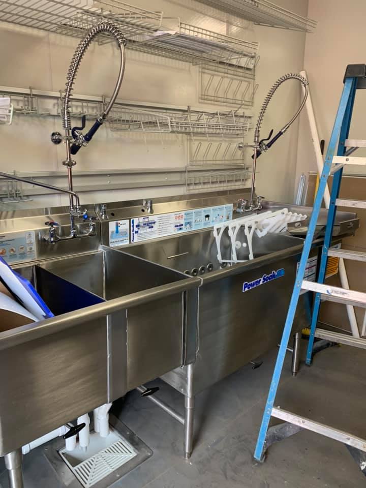 Stainless steel commercial sink with two spray faucets, surrounded by a ladder and wire shelving.